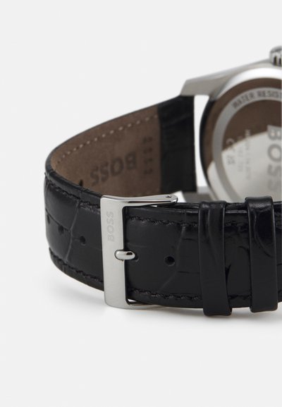 Bracelet de montre en cuir noir avec motif crocodile, fermoir en métal argenté avec gravure "BOSS", et dos en cuir marron ; couture détaillée.