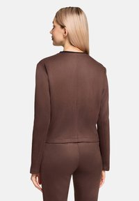 Brązowa dopasowana kurtka z długimi rękawami, gładką teksturą, okrągłym dekoltem i detalami szwu z tyłu. Noszona z pasującymi spodniami.
