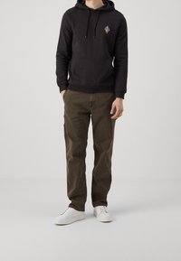 Felpa nera con tasca frontale e cordini, decorata con un piccolo logo colorato. I pantaloni verde oliva e le sneakers bianche completano l'outfit.