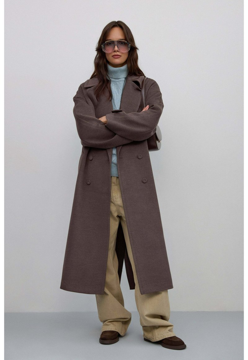 Brązowy długi płaszcz z wyciętym kołnierzem i podwójnym zapięciem, w połączeniu z jasnoniebieskim golfem, beżowymi szerokimi spodniami i brązowymi butami.