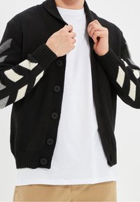 Cardigan nero in maglia con colletto a coste, dotato di grandi bottoni neri e maniche patterned con disegni zigzag in crema e grigio.