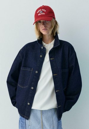 Mlada oseba z blond lasmi do ramen nosi rdečo kape "Costa Rica 1981 Golfito", temno modro denim jakno, belo polo majico in svetlo modre kavbojke.