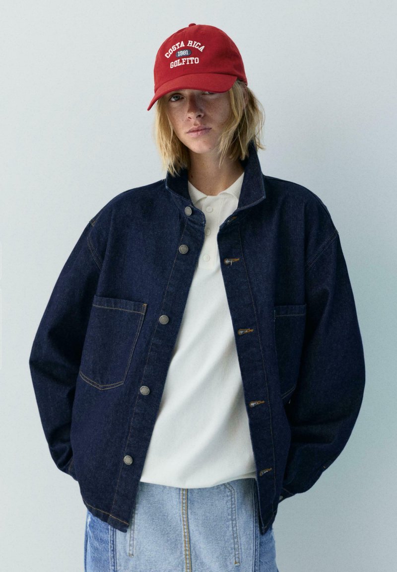 Jeune personne aux cheveux blonds mi-longs portant une casquette rouge "Costa Rica 1981 Golfito", une veste en jean foncé, un polo blanc et un jean bleu clair.