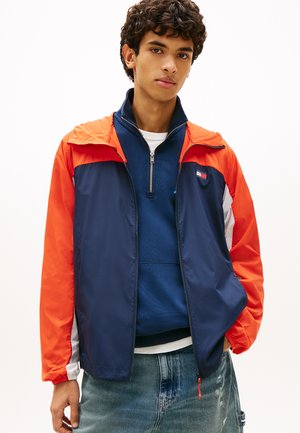 Mladý muž v námořnicky modrej a oranžovej vetrovke s farebnými blokmi, pod ktorou má námořnicky modrú mikinu so štvrťzipsom a vyblednuté modré džínsy.