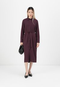 Woman in a purple buttoned midi dress with a waist tie, holding a black pouch bag and wearing black pointed heels.