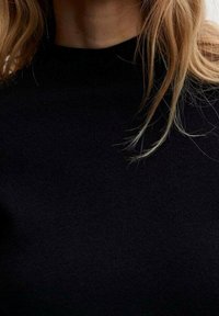 Pull noir en maille avec un col monté, texture lisse et silhouette ajustée. Des mèches de cheveux partiellement visibles au niveau du col.