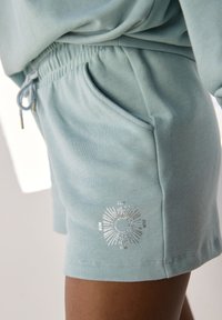 Gaiši zili kokvilnas šorti ar elastīgu jostasvietu, sānu kabatu un metālisku saules grafiku apakšmalā. Gluda tekstūra.