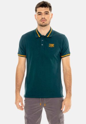 Jonge man die een donkergroen poloshirt draagt met oranje bies en "LEONE 1947" logo, gecombineerd met een grijze broek, staand voor een effen achtergrond.