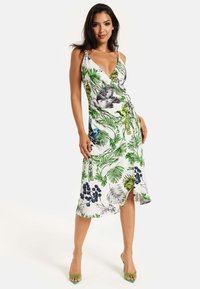 Tropical wrap dress in white fabric featuring green leaves, blue and purple flowers, and a thigh-high slit. Styled with green heels.