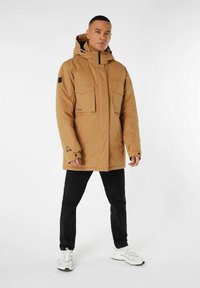 Braune Kapuzenjacke mit zwei Fronttaschen, verstellbaren Bündchen und einem Reißverschluss. Getragen über schwarzen Hosen und weißen Sneakers.