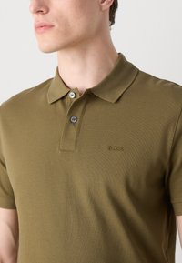 Tamsiai žalia polo marškinėliai, pagaminti iš tekstūruotos medžiagos, su apykakle, dviem mygtukais ir siuvinėtu "BOSS" logotipu krūtinėje.