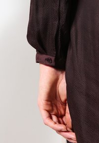 Zbliżenie na rękę spoczywającą obok, ubraną w ciemną, wzorzystą koszulę z długim rękawem i mankietem zapinanym na guziki.