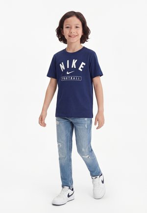 Niño sonriente con cabello hasta los hombros que lleva una camiseta de fútbol Nike azul marino, vaqueros desgarrados de color azul claro y zapatillas Nike blancas, de pie sobre fondo blanco.