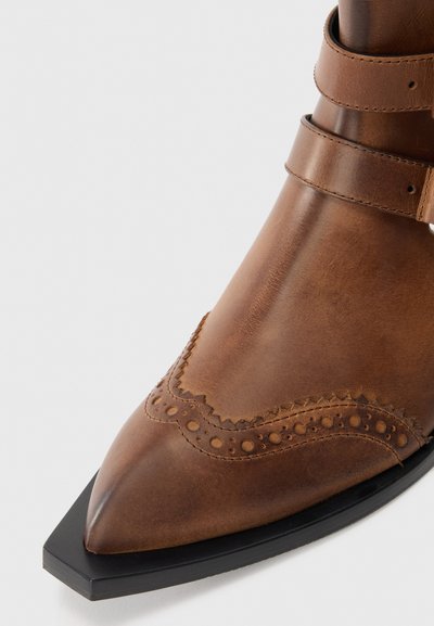 Botín de cuero marrón hasta el tobillo con punta afilada, perforaciones decorativas al estilo brogue y dos correas ajustables en la parte superior. Detalle de suela negra.