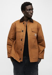 Νέος άνδρας που φοράει καφέ μπουφάν Carhartt πάνω από μαύρο πουκάμισο και σκούρα παντελόνια, στέκεται με τα χέρια στις τσέπες, κοιτάζοντας πλάγια.