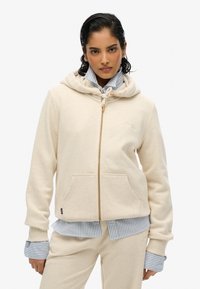 Veste à zip de couleur crème en tissu doux, dotée d'une capuche à cordon, de poches avant et de poignets côtelés, superposée sur une chemise rayée bleue.