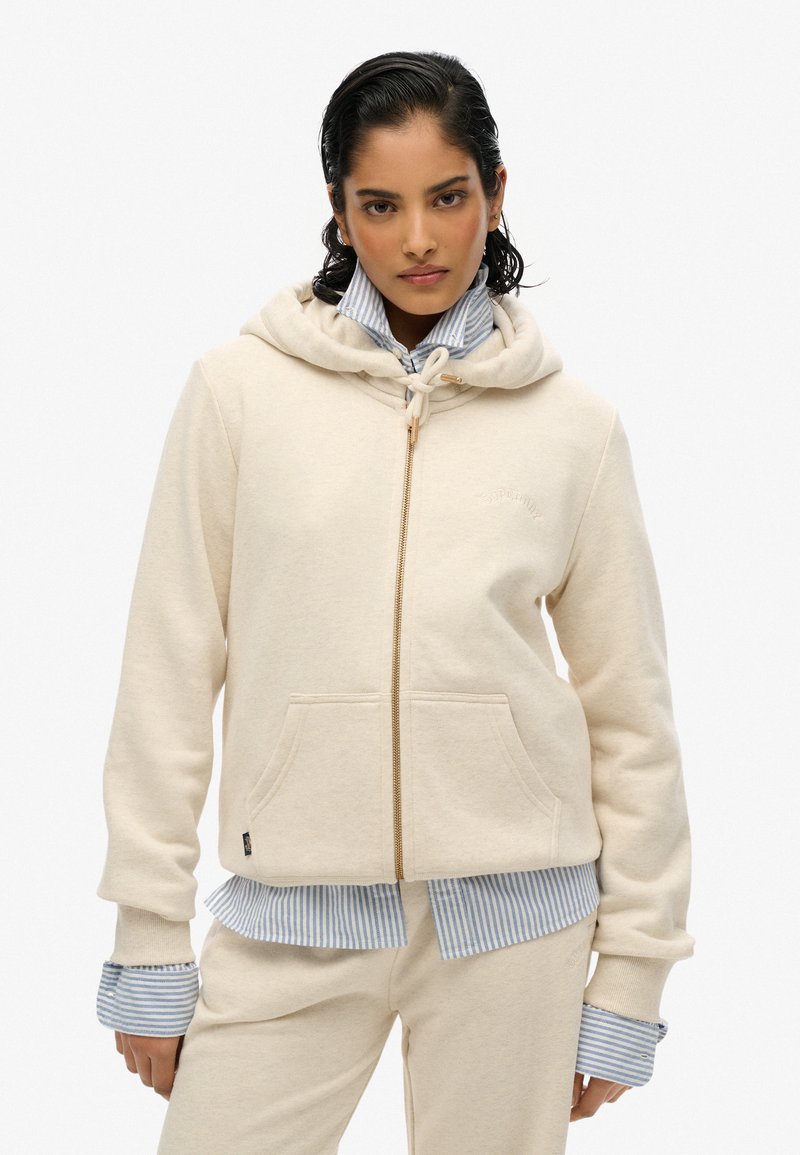Veste à zip de couleur crème en tissu doux, dotée d'une capuche à cordon, de poches avant et de poignets côtelés, superposée sur une chemise rayée bleue.