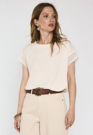 Donna che indossa una maglietta crema a maniche corte, pantaloni beige, cintura marrone e orecchini e braccialetti d'oro, guarda di lato.