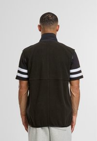 Gilet en polaire noir avec un col montant, sans manches. L'arrière présente une texture lisse et des lignes de couture subtiles. La chemise de dessus a des rayures blanches sur les manches.