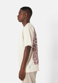 Kremowa koszulka z krótkim rękawem, z maroonowym wzorem graficznym na plecach. Gładka tekstura; prosty krój i luźny fit.