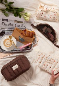 Borsa per cosmetici marrone trapuntata e pochette decorata con illustrazioni di cibo, accompagnate da una tazza di caffè, un croissant e un giornale.