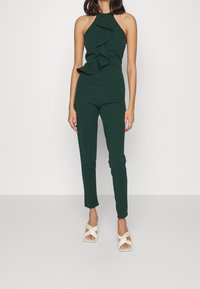 Mörkgrön jumpsuit med halterneck och volang fram, som har en figursydd silhuett och avsmalnade ben. Bärs med vita sandaletter med remmar.