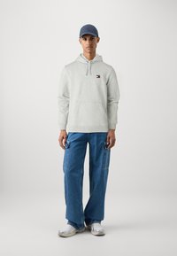 Sudadera gris con bolsillo frontal y detalle de logo, combinada con unos vaqueros azules de corte holgado que tienen bolsillos tipo cargo. Zapatillas blancas completan el conjunto.
