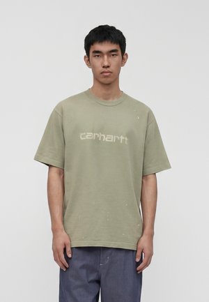 Junger Mann mit kurzen schwarzen Haaren, trägt ein ausgewaschenes olivgrünes Carhartt T-Shirt und lockere dunkelblaue Hose vor einfarbigem Hintergrund.