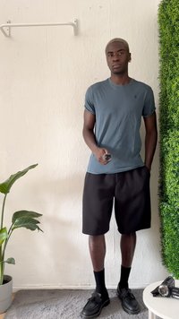 Μοντέλο φοράει μπλε κοντομάνικο T-shirt και μαύρες βερμούδες. Μαύρα παπούτσια και κάλτσες. Το φόντο περιλαμβάνει ένα πράσινο φυτό και έναν υφασμάτινο τοίχο.