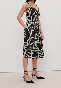 Robe à motifs noir et crème avec un corsage ajusté, un décolleté en V, des poches latérales et une jupe évasée. Associée à des escarpins noirs à talons.