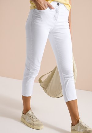 Schmal geschnittene weiße Caprihose getragen mit beigen Sneakers, einem gelben Oberteil und einer beigen Tasche in der linken Hand.