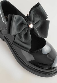Chaussure en cuir verni noir avec un large nœud en ruban noir à côtes et un accent en cœur en or rose. Bout arrondi et semelle noire texturée.