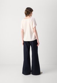 Beige top s krátkým rukávem a čtvercovým výstřihem, spárovaný s tmavě modrými rozšířenými džíny. Hladká textura, volný střih a viditelné detaily šití.