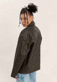 Barna műbőr dzseki széles gallérral és látható varrási részletekkel, világoskék farmernadrággal párosítva.