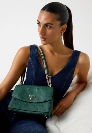 Sac à main en cuir vert texturé avec un rabat, une chaîne en bandoulière et des détails de logo à l'avant. Positionné à côté d'un modèle portant du denim.