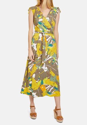 Donna che indossa un vestito midi senza maniche con cintura, con una fantasia colorata di foglie tropicali e sandali con zeppa marroni, in piedi su sfondo bianco.