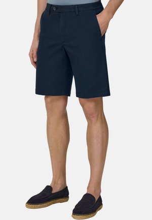 Mężczyzna w granatowych krótkich spodenkach do kolan z bocznymi kieszeniami oraz czarnych wsuwanych butach z tkanymi podeszwami, stojący na jednolitym tle.