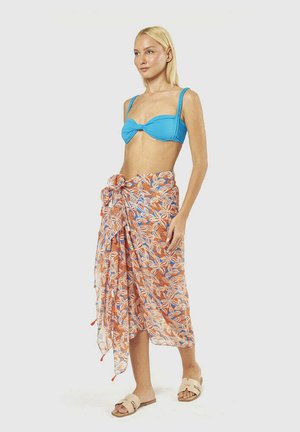 Donna con top bikini blu che indossa un sarong floreale arancione e bianco legato in vita e sandali beige, in piedi contro uno sfondo semplice.