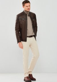 Brown leather jacket with a high collar, zip pockets, and embossed logos, worn over a beige turtleneck and light-colored trousers.