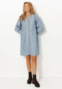 Robe en denim bleu clair à manches longues, avec un devant à boutons et une broderie blanche complexe. Bottines brunes à la cheville avec une semelle épaisse.