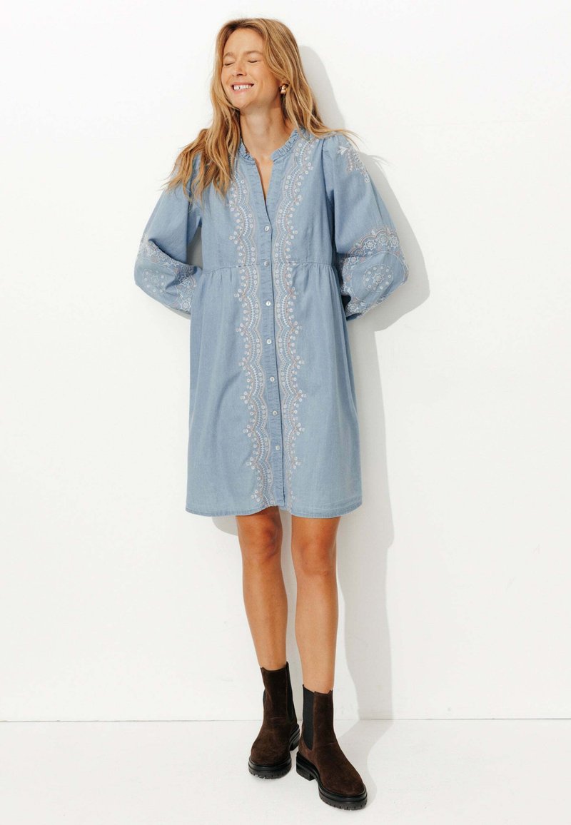 Robe en denim bleu clair à manches longues, avec un devant à boutons et une broderie blanche complexe. Bottines brunes à la cheville avec une semelle épaisse.