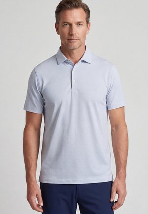 Mann trägt ein hellblaues kurzärmeliges Poloshirt mit dunkelblauer Hose und steht vor einem schlichten grauen Hintergrund.