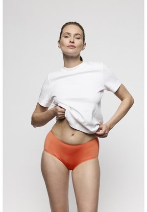 Vrouw tilt wit T-shirt op om oranje high-waist ondergoed te laten zien tegen een effen lichtgrijze achtergrond.