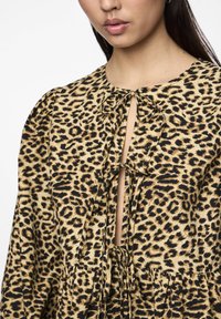 Μπλούζα λεοπάρ με σχέδιο που διαθέτει λαιμόκοψη τύπου κλειδαριά, λεπτομέρειες με κόμπους και τριπλή σχεδίαση. Το ύφασμα έχει λεία υφή με αντίθετες μαύρες και μπεζ αποχρώσεις.