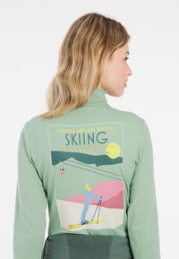 Jasnozielona, długorękawowa góra narciarska z graficznym nadrukiem narciarza, gór i gondoli na tle w pastelowych kolorach.