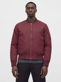 Homme portant une veste bomber bordeaux zippée avec poignets et ourlet côtelés, debout devant un fond gris clair uni.