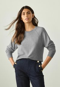Grå sweatshirt med rund hals og ribbede manchetter, parret med mørke denimbukser med guldknapdetaljer på lommerne.
