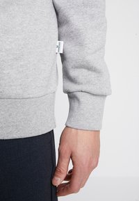 Tessuto grigio della felpa con polsini a coste, che mostra una mano appoggiata su pantaloni scuri. Piccolo etichetta del marchio visibile sulla cucitura laterale della felpa.