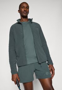 Ciemnozielona polarowa kurtka z zamkiem, noszona na teksturowanej ciemnozielonej koszuli i dopasowanych szortach. Nosi subtelną markową metkę.