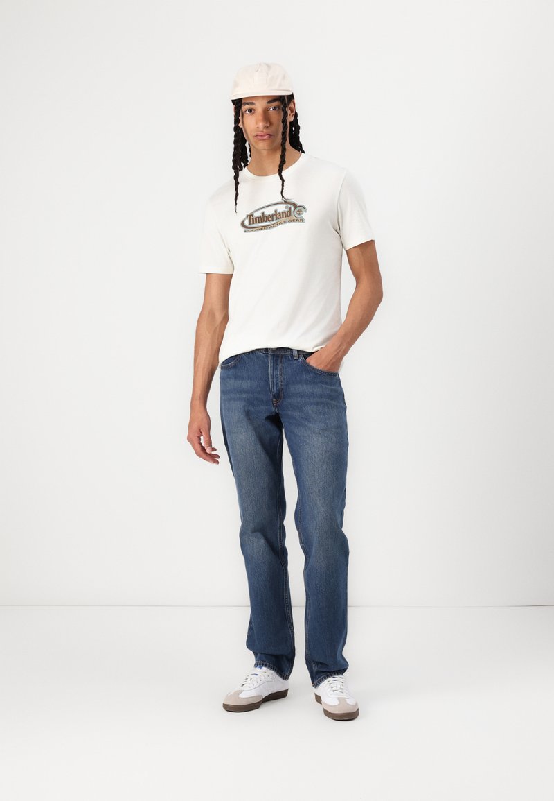 Witte t-shirt met "Timberland" logo, blauwe spijkerbroek en beige pet. Wit-grijze sneakers met textuurdetails. Neutrale achtergrond.